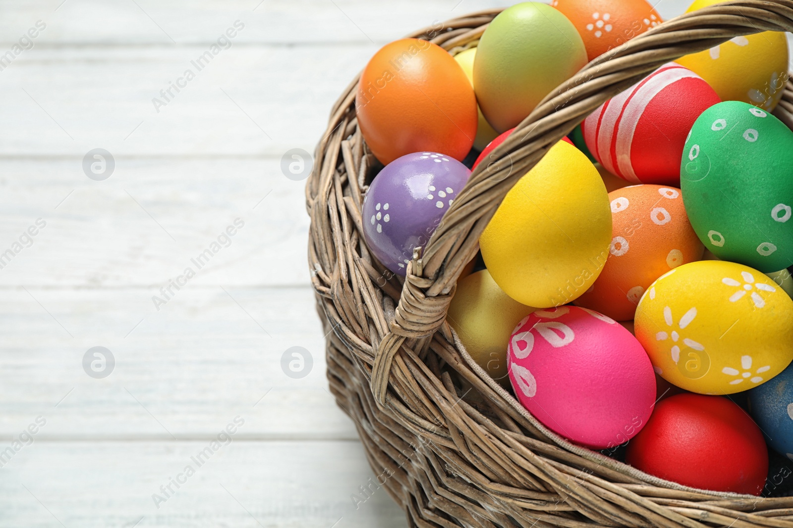 Photo of Colorful Easter eggs in basket on white wooden table, closeup. Space for text