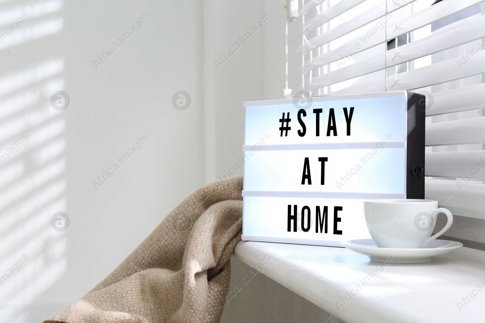 Photo of Cup of drink and lightbox with hashtag STAY AT HOME on window sill indoors. Message to promote self-isolation during COVID‑19 pandemic