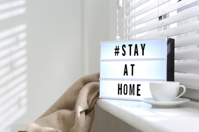 Cup of drink and lightbox with hashtag STAY AT HOME on window sill indoors. Message to promote self-isolation during COVID‑19 pandemic