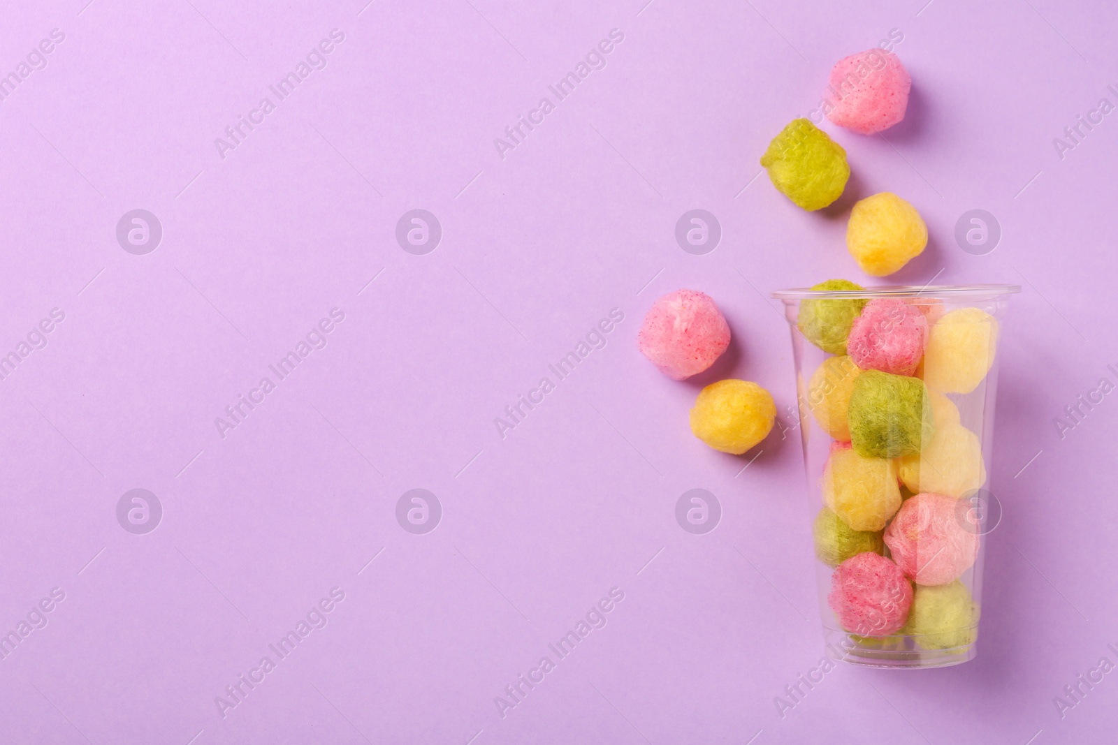 Photo of Plastic cup with color cotton balls on violet background, flat lay and space for text. Sweet candy