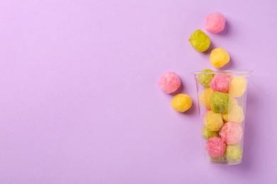 Photo of Plastic cup with color cotton balls on violet background, flat lay and space for text. Sweet candy