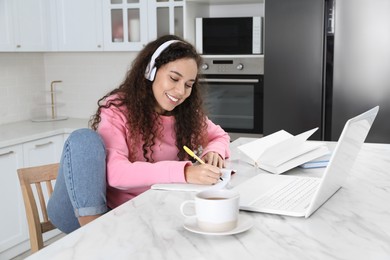 Photo of African American woman with modern laptop and headphones studying in kitchen. Distance learning
