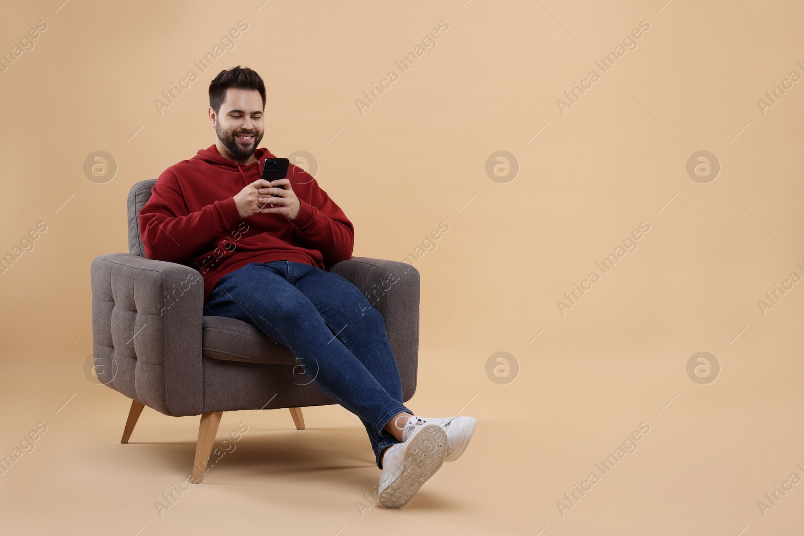 Photo of Happy young man using smartphone on armchair against beige background, space for text