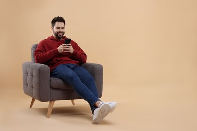 Happy young man using smartphone on armchair against beige background, space for text