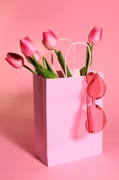 Photo of Shopping bag with beautiful tulips and stylish sunglasses on pink background