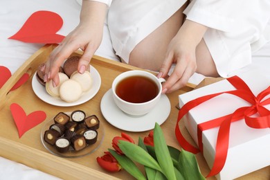 Photo of Tasty breakfast served in bed. Woman with tea, desserts, gift box and flowers at home, closeup