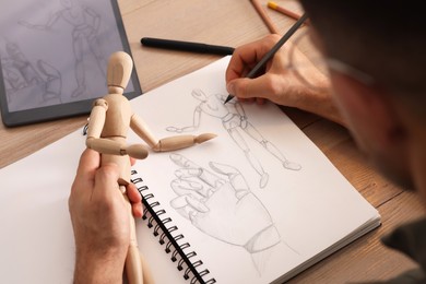 Man drawing mannequin in sketchbook with pencil at wooden table, closeup