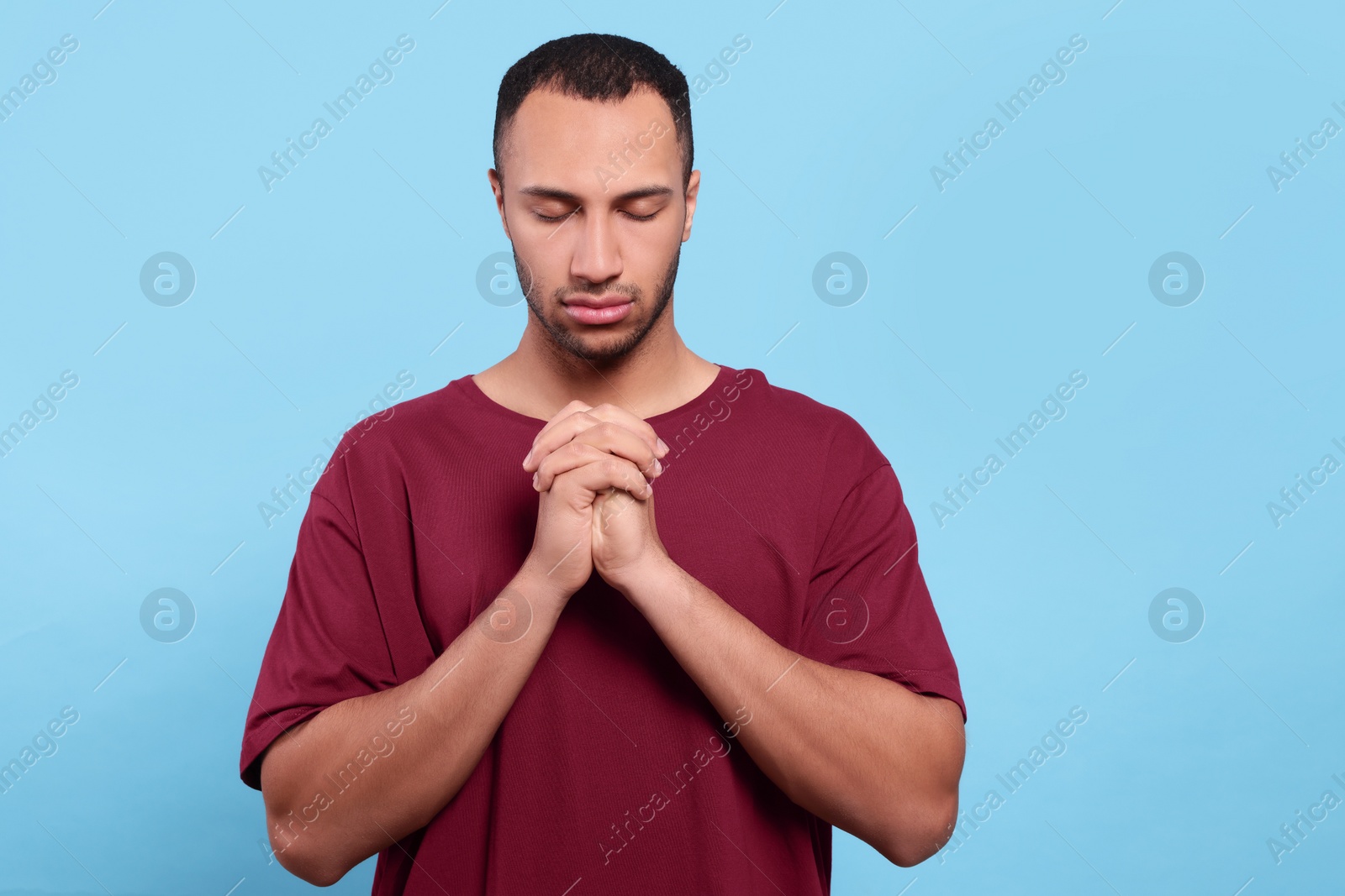 Photo of African American man with clasped hands praying to God on light blue background