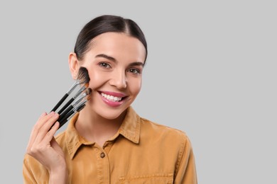 Happy woman with different makeup brushes on light grey background, space for text