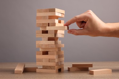 Photo of Playing Jenga. Man removing block from tower at wooden table against grey background, closeup