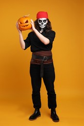 Photo of Man in scary pirate costume with skull makeup and carved pumpkin on orange background. Halloween celebration