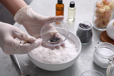 Photo of Woman in gloves filling bath bomb mold with dried flower buds at grey table, closeup