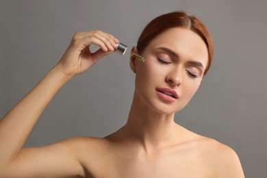 Beautiful young woman applying cosmetic serum onto her face on grey background