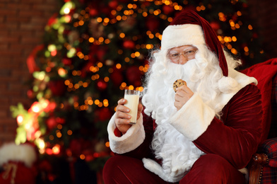 Photo of Santa Claus with milk and cookie near Christmas tree indoors