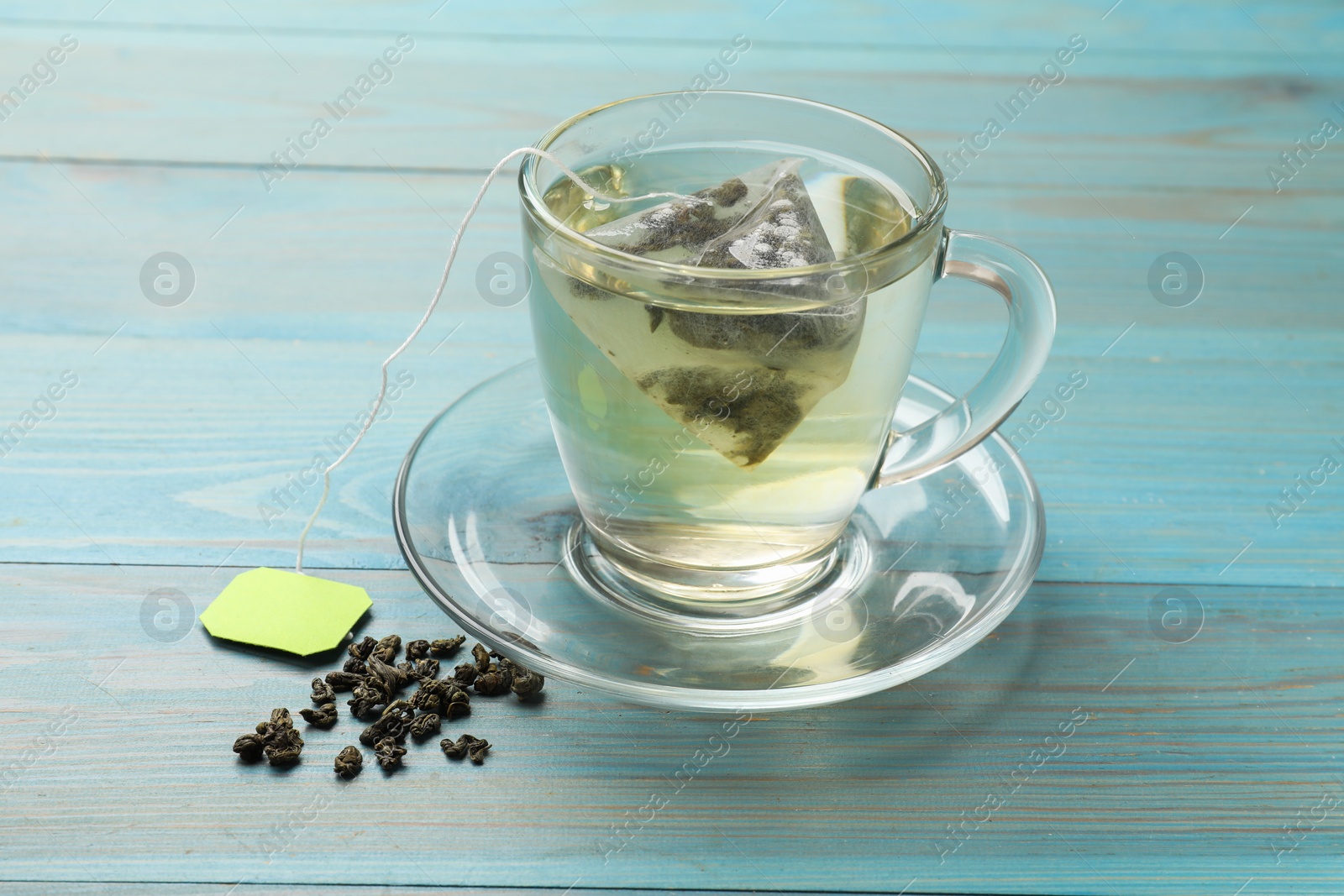 Photo of Tea bag in cup with hot drink and dry leaves on light blue wooden table