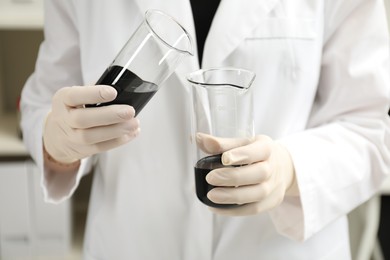Photo of Laboratory worker pouring black crude oil into beaker indoors, closeup