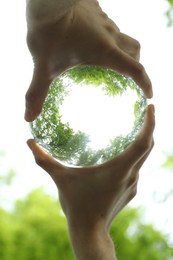 Overturned reflection of beautiful green trees outdoors, low angle view. Man holding crystal ball in park, closeup