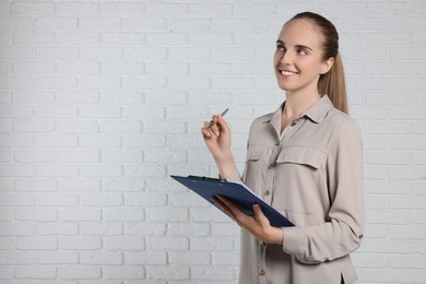 Photo of Happy young secretary with clipboard and pen near white brick wall, space for text