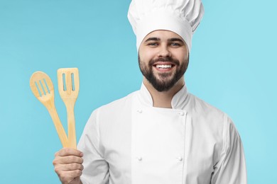 Photo of Happy young chef in uniform holding wooden utensils on light blue background