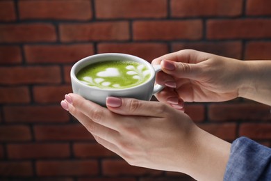 Photo of Woman with cup of delicious matcha latte against brick wall, closeup