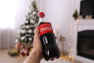 MYKOLAIV, UKRAINE - JANUARY 13, 2021: Woman holding bottle of Coca-Cola in room with Christmas tree, closeup