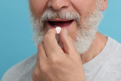 Senior man taking pill on light blue background, closeup