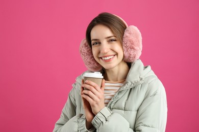 Happy woman with cup of drink wearing warm earmuffs on pink background