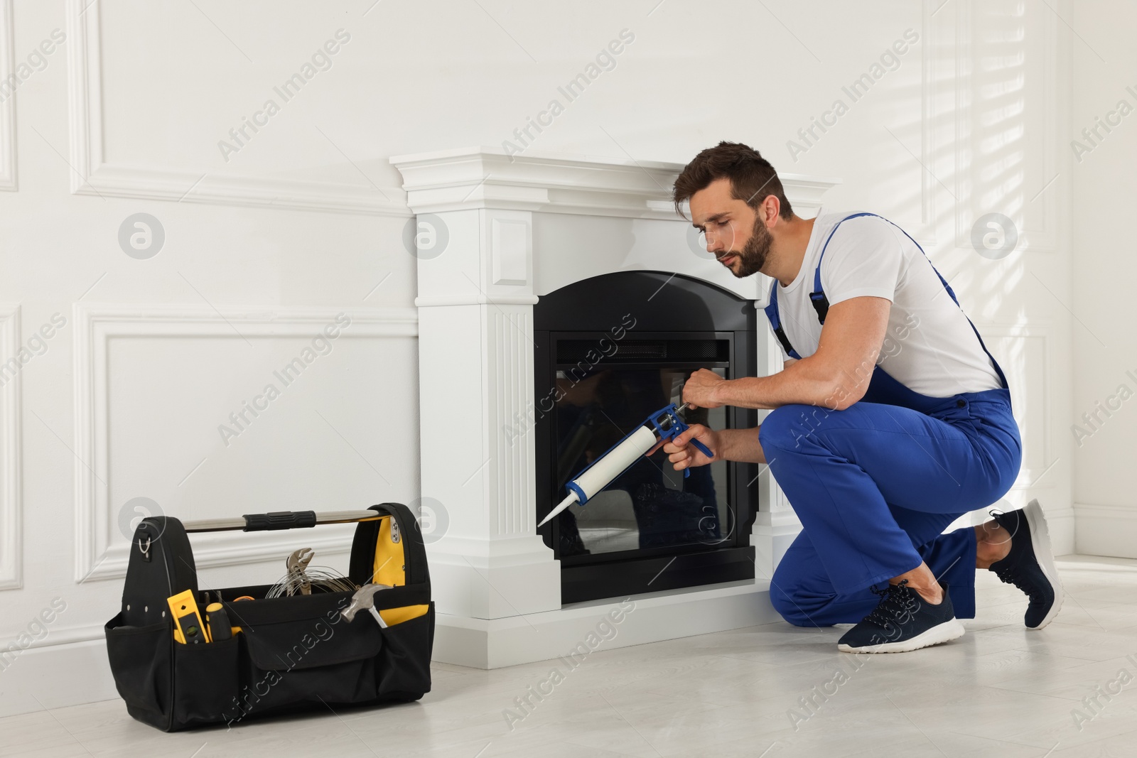 Photo of Professional technician sealing electric fireplace with caulk near white wall in room