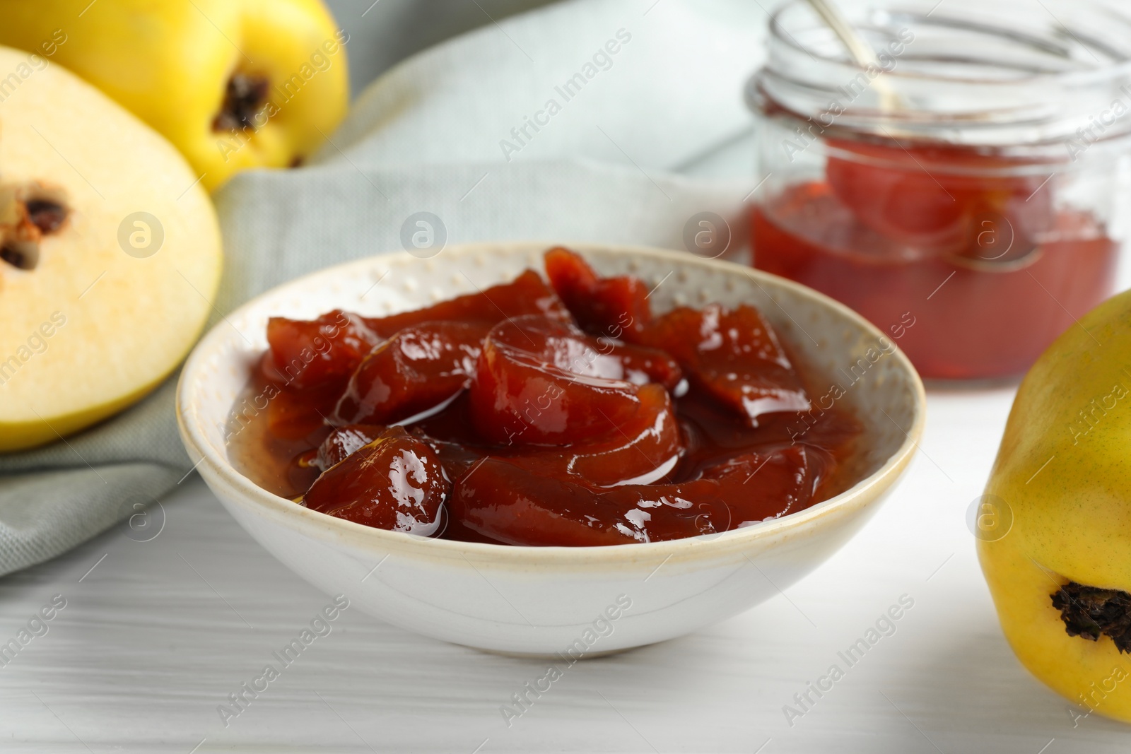 Photo of Tasty homemade quince jam in bowl and fruits on white wooden table, closeup