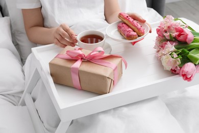 Photo of Tasty breakfast served in bed. Woman with tea, eclairs, gift box and flowers at home, closeup