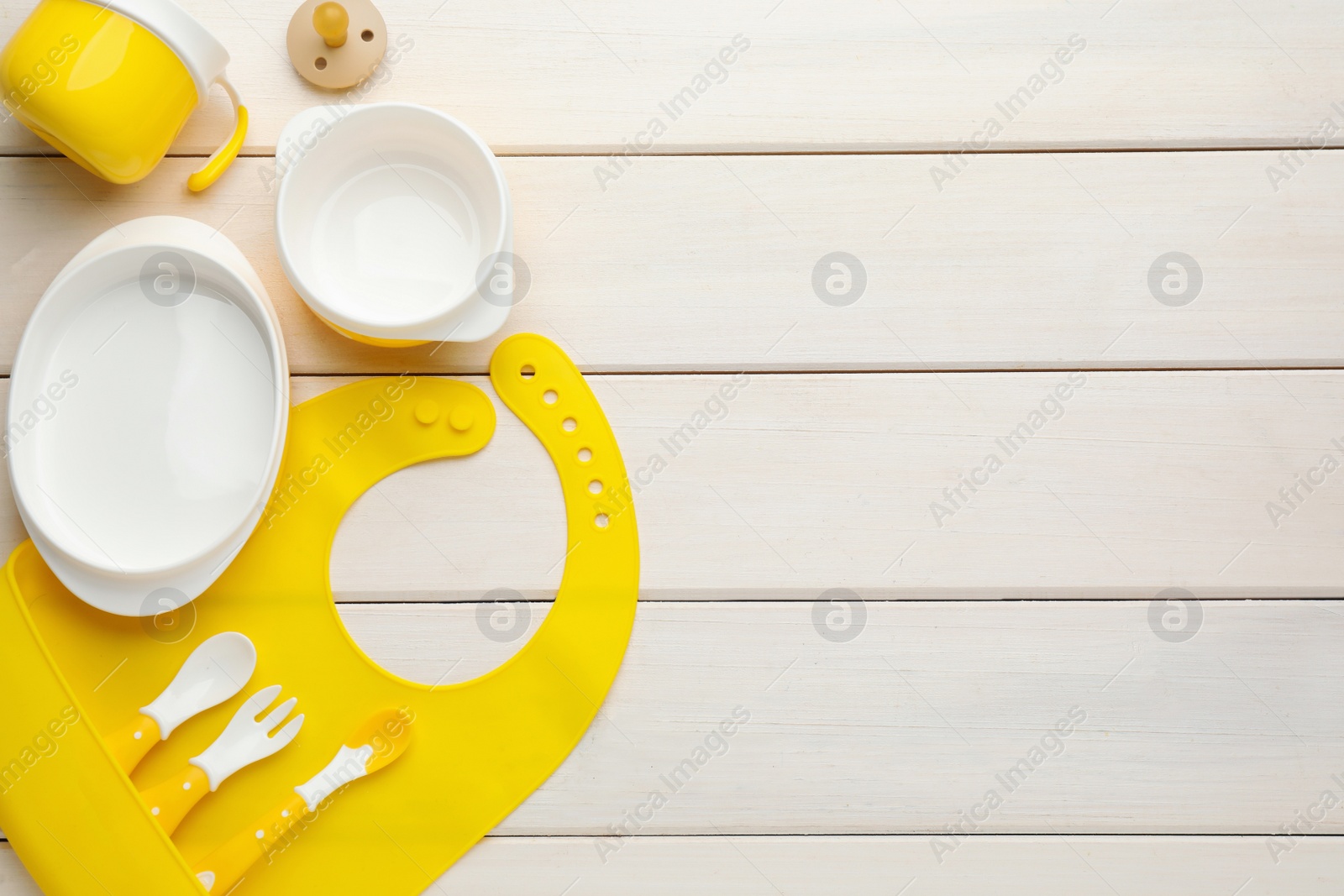 Photo of Flat lay composition with silicone baby bib and plastic dishware on white wooden background. Space for text