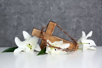 Photo of Wooden cross, crown of thorns and blossom lilies on table against color background