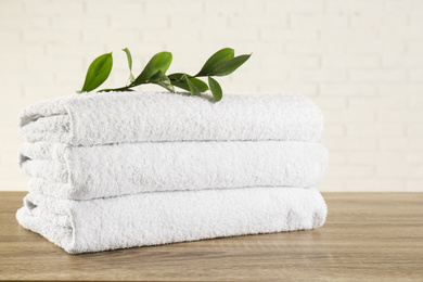 Photo of Stack of clean bath towels and green branch on wooden table near white brick wall
