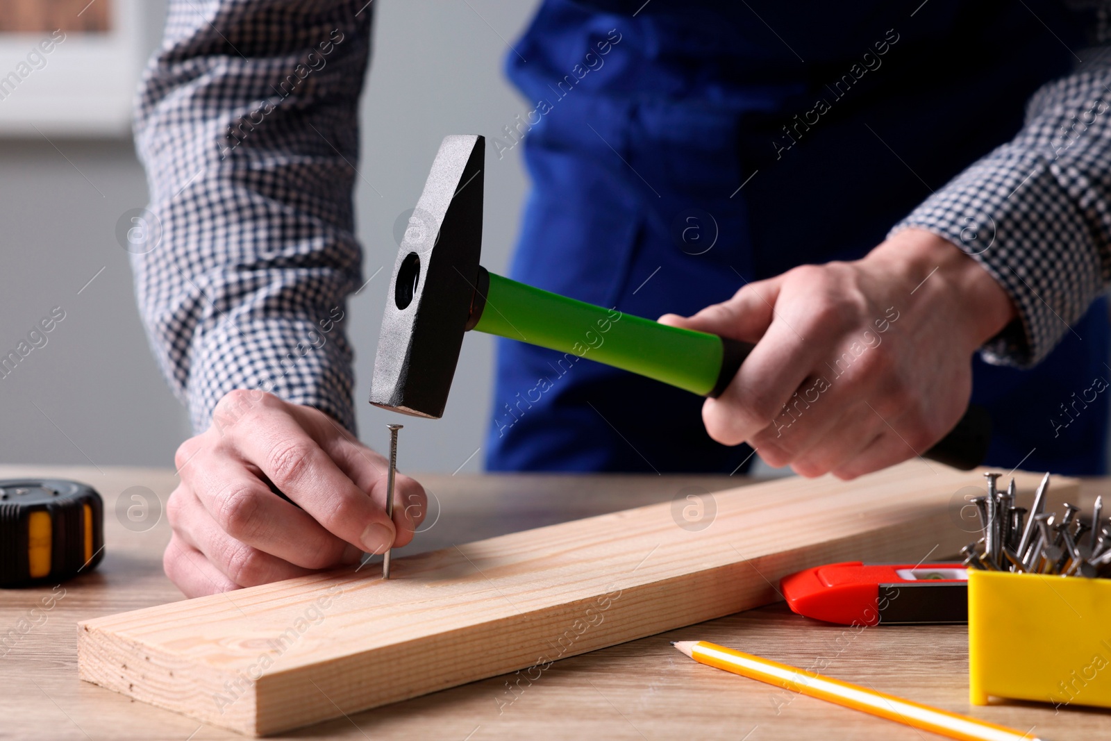 Photo of Professional repairman hammering nail into board at wooden table indoors, closeup