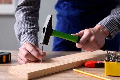 Photo of Professional repairman hammering nail into board at wooden table indoors, closeup