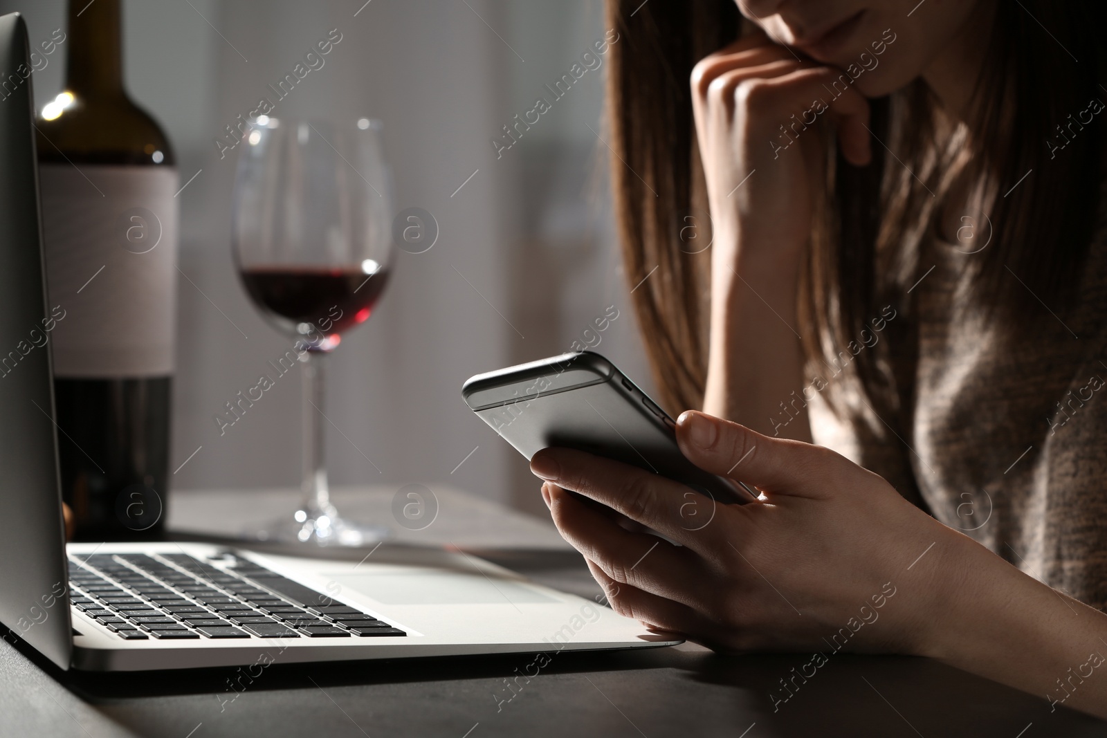 Photo of Woman using smartphone at table with laptop near glass of wine indoors, closeup. Loneliness concept