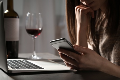 Photo of Woman using smartphone at table with laptop near glass of wine indoors, closeup. Loneliness concept