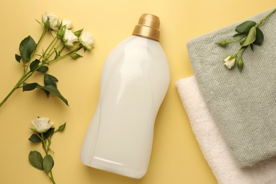 Photo of Bottle of fabric softener, terry towels and roses on pale yellow background, flat lay