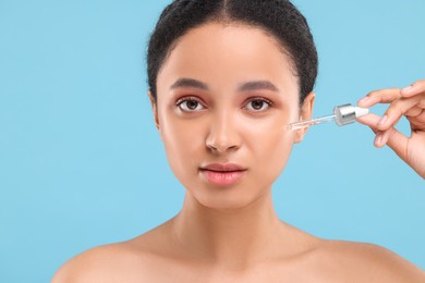 Beautiful woman applying serum onto her face on light blue background
