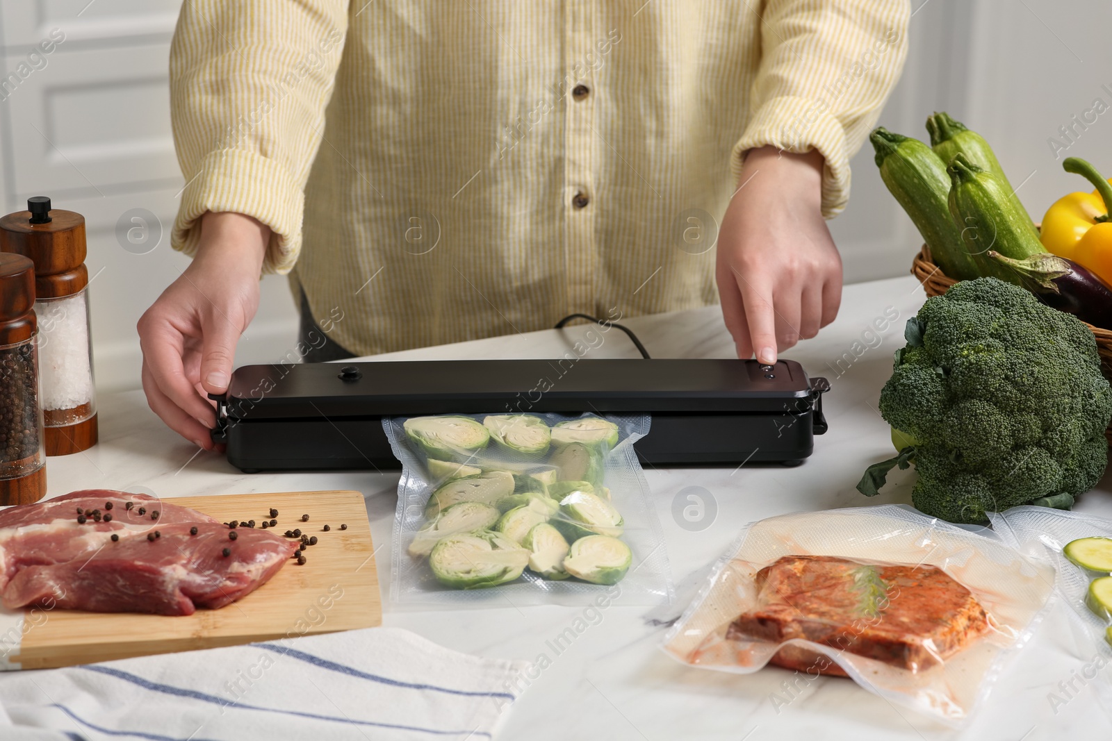 Photo of Woman using sealer for vacuum packing with plastic bag of zucchini at white table in kitchen, closeup