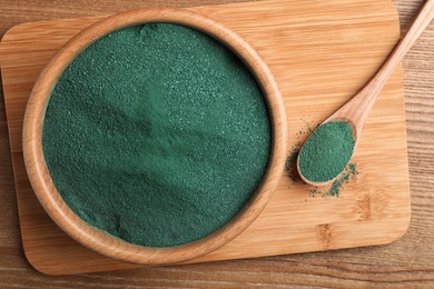 Photo of Board with bowl and spoon of spirulina powder on wooden background, top view