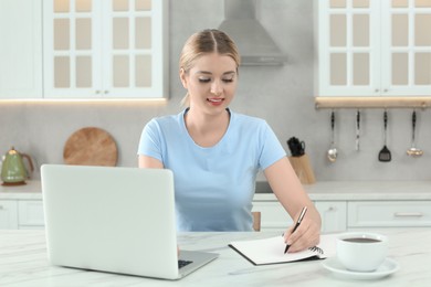 Photo of Home workplace. Woman writing in notebook near laptop at marble desk in kitchen