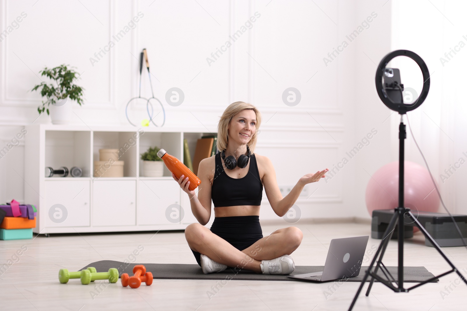 Photo of Smiling sports blogger holding bottle while streaming online fitness lesson at home