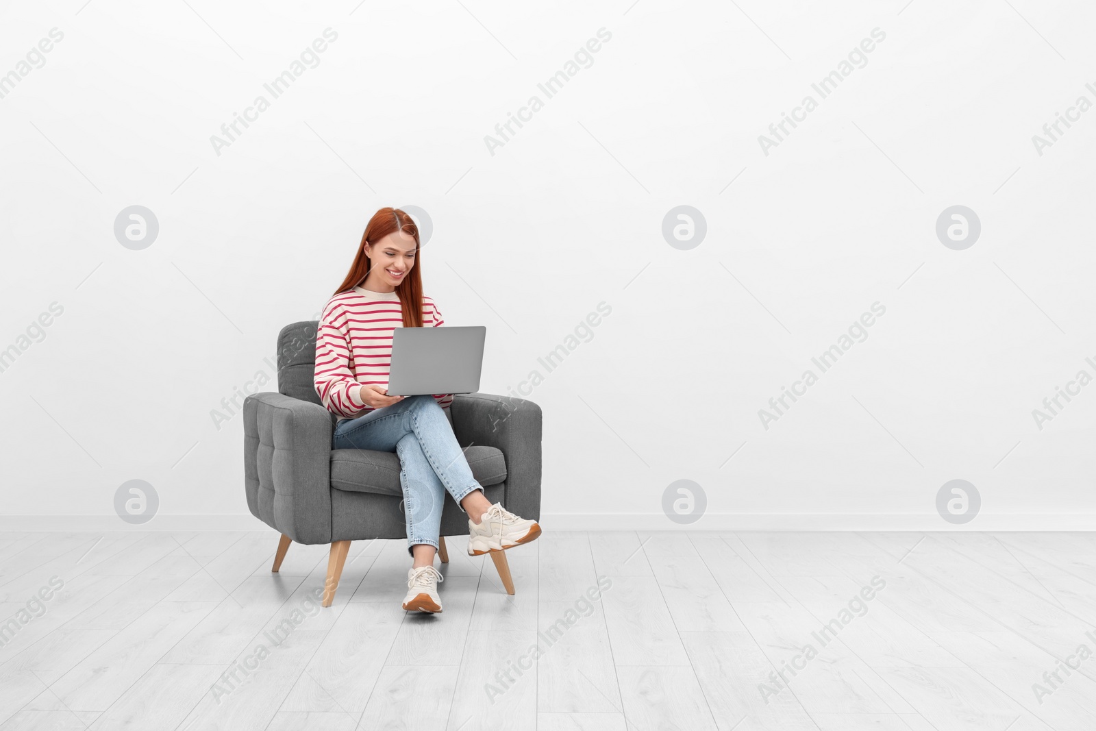 Photo of Happy young woman with laptop sitting in armchair near white wall indoors. Space for text