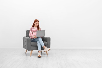 Happy young woman with laptop sitting in armchair near white wall indoors. Space for text