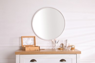 Trendy round mirror and chest of drawers with accessories near white wall indoors.