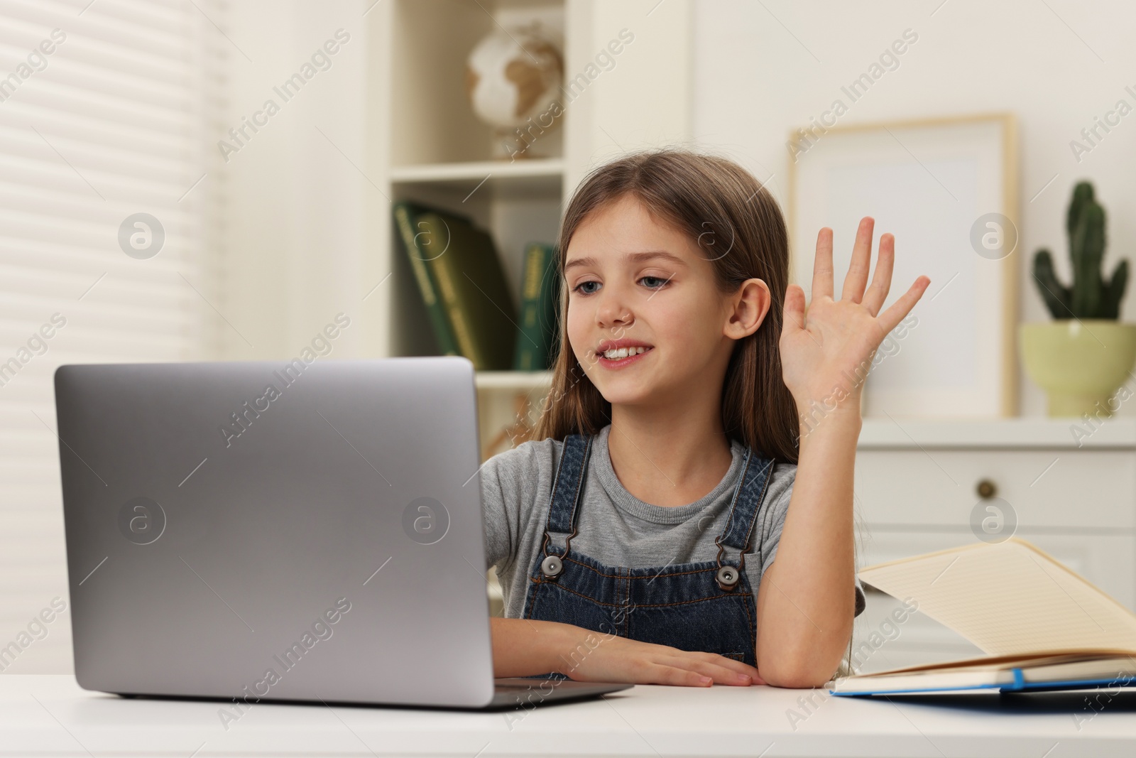 Photo of Cute girl waving hello during online lesson via laptop at white table indoors