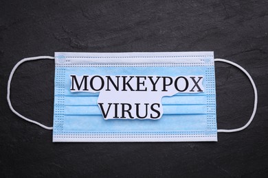 Medical face mask with words Monkeypox Virus on dark background, top view
