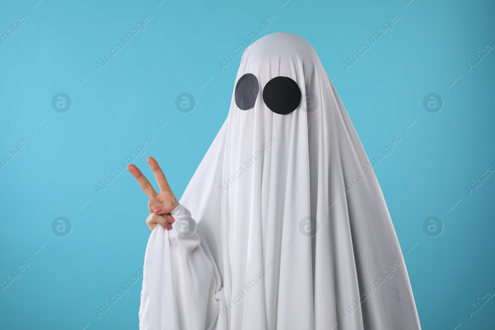 Photo of Funny ghost. Woman covered with white sheet showing V-sign on light blue background