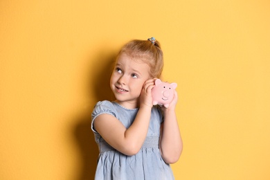 Little girl with piggy bank on color background